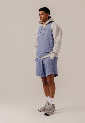 Jeune homme debout, les mains dans les poches, portant un sweat à capuche beige sous un gilet bleu à motifs et un short assorti, avec des chaussettes blanches et des baskets.