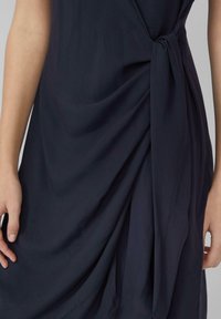 Dunkelblaues Kleid mit verdrehtem Vorderteil, aus leichtem Stoff, mit sanften Drapierungen und strukturiertem Finish. Mit einem Knoten-Detail akzentuiert.