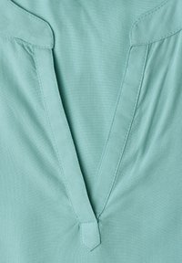Gros plan sur un tissu vert clair avec une encolure en V cousue et un petit détail de languette en tissu à la base.
