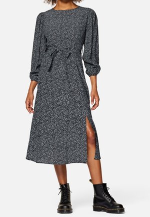 Mittelanges Kleid mit Blumenmuster in Marineblau und Weiß, mit Rundhalsausschnitt, Puffärmeln, Taillenschlaufe und Seitenschlitz. Kombiniert mit schwarzen Stiefeln.