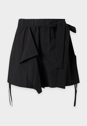 Jeune homme debout portant une chemise à manches longues gris clair, un short noir noué avec tissu superposé, des chaussettes blanches et des mocassins noirs sur fond blanc.