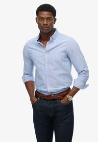 Jasnoniebieska koszula z guzikami w różowe paski, dopasowany krój, podwinięte rękawy. Połączona z ciemnymi dżinsami i plecionym brązowym paskiem.