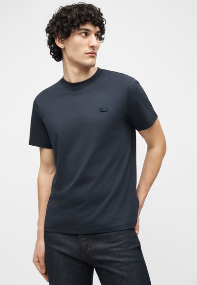 Jeune homme aux cheveux bouclés portant un t-shirt bleu marine foncé et un jean sombre, regardant vers sa gauche sur un fond clair uni.