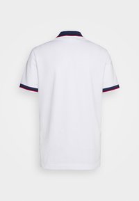 Polo blanc à manches courtes avec col bleu marine et bords des manches, chacun souligné par un fin liseré rouge, vu de dos.