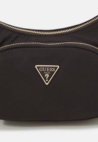 Černá nylonová kabelka s kulatým tvarem, zlatými kovovými doplňky včetně zipů a trojúhelníkovou plaketou s logem „GUESS“ a otazníkem.