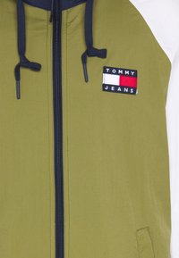 Giacca a zip verde oliva con colletto blu navy, caratterizzata da un logo a patch nero con accenti rossi, bianchi e blu. Texture liscia, interno foderato.