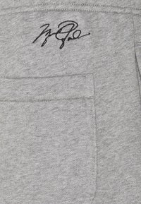Tejido de sudadera gris con una textura suave; presenta un bolsillo y una firma bordada en negro en la zona del pecho.