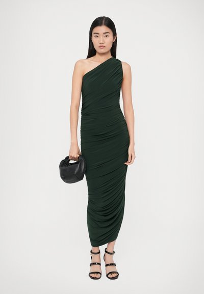 Tmavozelené asymetrické šaty s textúrou s nariaseným efektom, priliehavého strihu. Modelka drží čiernu kabelku a nosí čierne sandále na remienky.