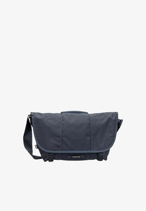 Borsa messenger Timbuk2 blu scuro con tracolla regolabile, patta frontale e due fibbie nere su sfondo bianco.