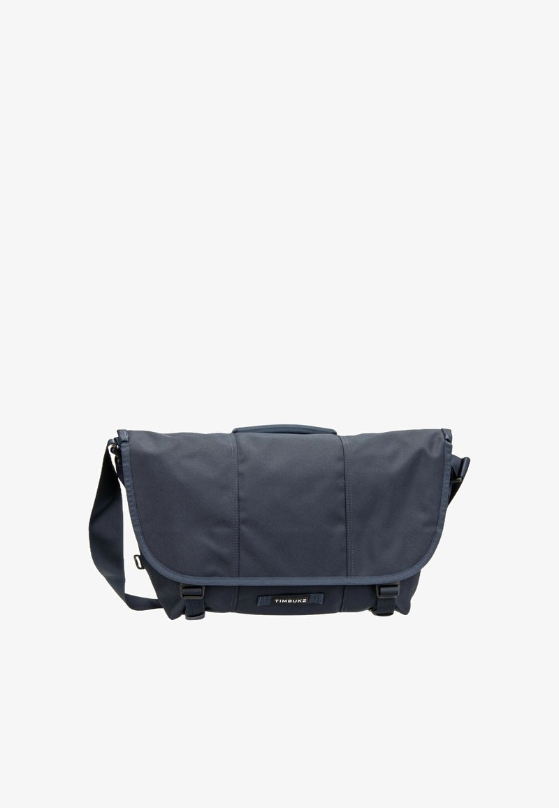 Sac messager Timbuk2 bleu foncé avec bandoulière réglable, rabat avant et deux fermetures à boucles noires sur fond blanc.