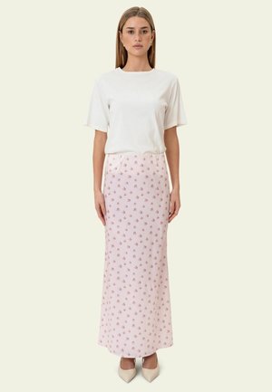 Kobieta o prostych włosach nosi biały krótki top, jasnoróżową spódnicę do kostek z drobnym kwiatowym wzorem oraz beżowe buty na szpic, stoi nieruchomo.