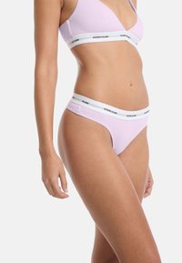 Lichtpaarse bralette en bijpassende thongonderbroek met een witte elastische tailleband met de branding "GUESS JEANS". Glad stoftextuur.