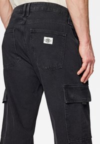 Pantaloni cargo neri con molte tasche, tessuto denim testurizzato e una piccola etichetta bianca con il marchio sul retro. La cintura presenta passanti per la cintura.