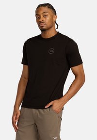 T-shirt nera a maniche corte realizzata in tessuto liscio, con un logo discreto sul petto, abbinata a pantaloni corti marrone chiaro.
