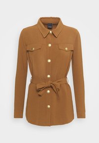 Giacca marrone su misura in un tessuto morbido, con chiusura a bottoni, due tasche sul petto e un dettaglio in vita annodato con bottoni color oro.