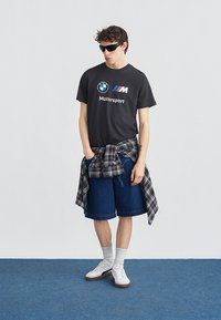 Νεαρός άνδρας με μαύρο μπλουζάκι BMW Motorsport, καρό πουκάμισο δεμένο γύρω από τη μέση, τζιν σορτς, λευκά αθλητικά παπούτσια και σκούρα γυαλιά ηλίου στέκεται σε μπλε χαλί.