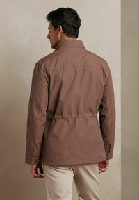 Brązowa, lekka kurtka z kołnierzem stójkowym, elastycznym pasem i mankietami zapinanymi na guziki. Gładka tekstura i minimalistyczny design.