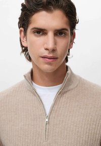 Maglione beige con zip, texture a coste, colletto alto e una t-shirt bianca sotto, che presenta un design rilassato e semplice.
