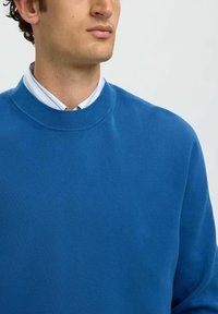 Homme portant un pull bleu à col rond par-dessus une chemise blanche à col avec des rayures bleues, sur un fond clair uni.