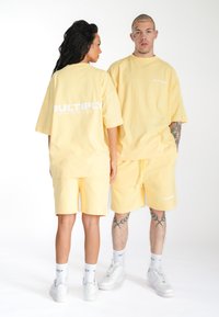 Gule oversized bomulds T-shirts og shorts. T-shirts har "MULTIPLY" tryk på ryggen, parret med hvide sokker og sneakers.