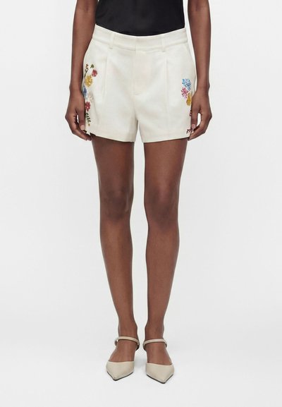 Femme portant un short crème avec une broderie florale colorée de chaque côté, un haut noir et des chaussures crème à bout pointu avec brides à la cheville.