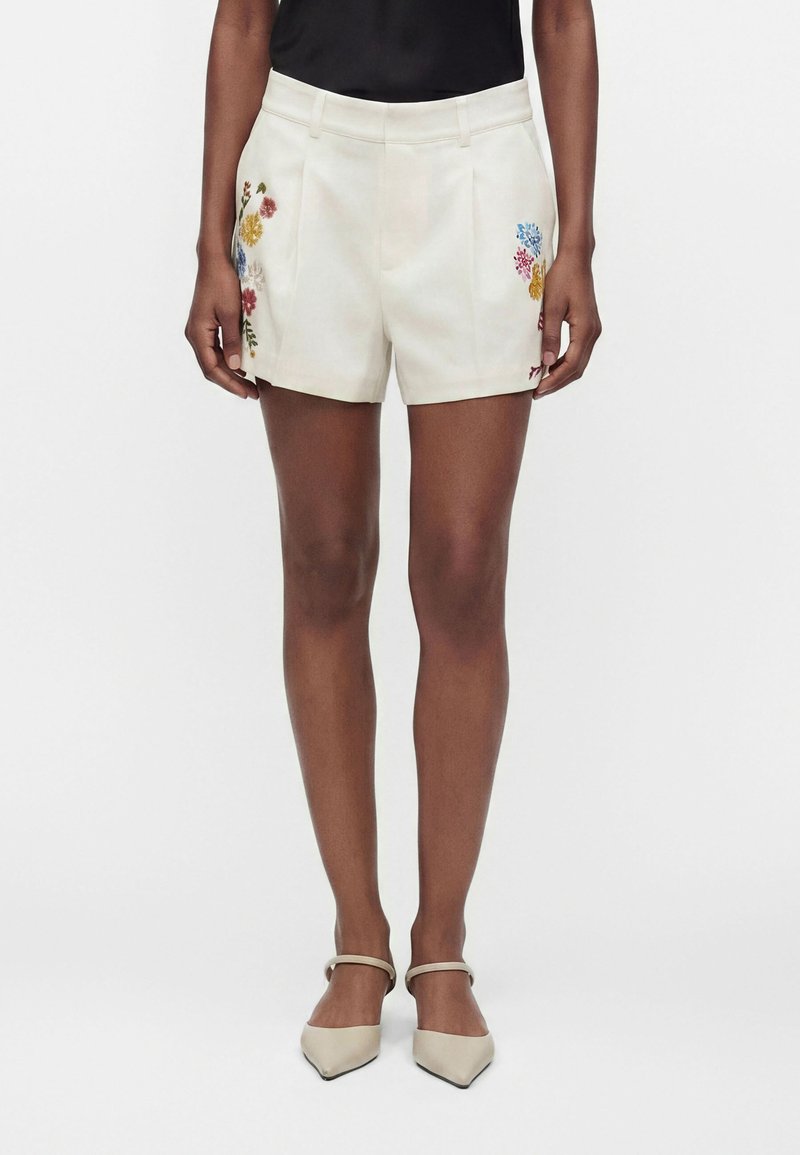 Femme portant un short crème avec une broderie florale colorée de chaque côté, un haut noir et des chaussures crème à bout pointu avec brides à la cheville.