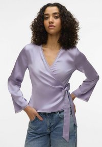 Top croisé en lavande avec des manches longues, finition en dentelle et lien sur le côté. Tissu lisse et léger ; associé à un jean taille haute.