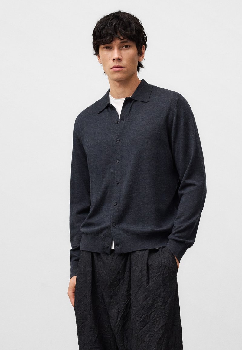 Cardigan grigio scuro con colletto, chiusura a bottoni e polsini a coste. Indossato su una maglietta bianca, abbinato a pantaloni neri strutturati.