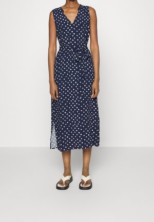 Femme portant une robe midi bleu marine sans manches avec un motif losange blanc et une ceinture à la taille, associée à des sandales à plateforme blanches.