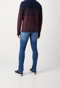 Persona vestida con un suéter a rayas en tonos azul marino y burdeos, jeans ajustados azules y zapatillas negras de corte bajo, de pie de espaldas a la cámara.