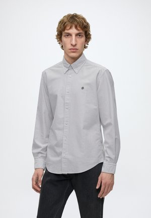 Jeune homme portant une chemise à rayures gris clair à boutons avec manches longues et pantalon sombre, debout devant un fond blanc uni.