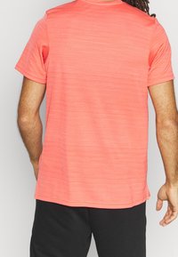 Camiseta de manga corta color coral en tejido texturizado, con cuello acanalado y dobladillo recto, llevada con pantalones cortos negros, vista desde la parte de atrás.