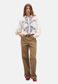 White blouse with blue floral embroidery, puffed sleeves, and black trim; paired with tan straight-leg pants and red shoes.