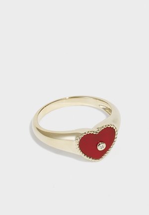 Gold-tone ring featuring a red heart-shaped detail with a small gemstone in the center, outlined with a textured gold border.