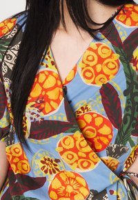 Haut cache-cœur floral avec un fond bleu et des motifs vibrants de fleurs et feuilles en orange, jaune et vert. Tissu lisse, manches courtes.