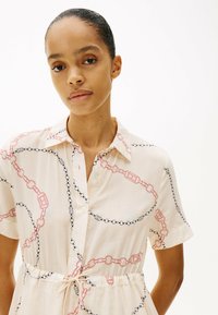 Kortærmet kjole i lysebeige med et kædelinkmønster i rød og sort. Har krave og et bindebånd i taljen.