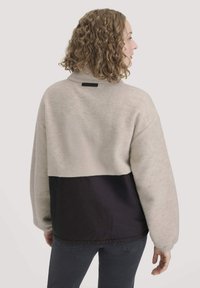 Fleecepullover mit einem cremefarbenen oberen Abschnitt und einem dunkelmarineblauen unteren Teil, mit hohem Kragen und einem rechteckigen schwarzen Etikett am Hals.