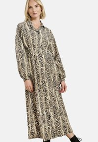 Žena v dlouhém rukávu, v šatech s potiskem hada a límečkem, s předními knoflíky a černými kotníkovými botami, stojící na bílém pozadí.