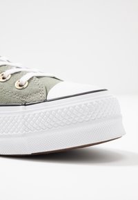 Green canvas sneaker with a white rubber toe cap and textured white sole. Features white laces and gold eyelets for fastening.