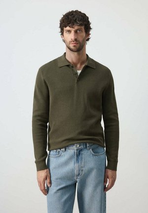 Hombre con cabello rizado y oscuro que lleva una camisa polo verde oliva de manga larga y jeans azul claro, de pie frente a un fondo liso.