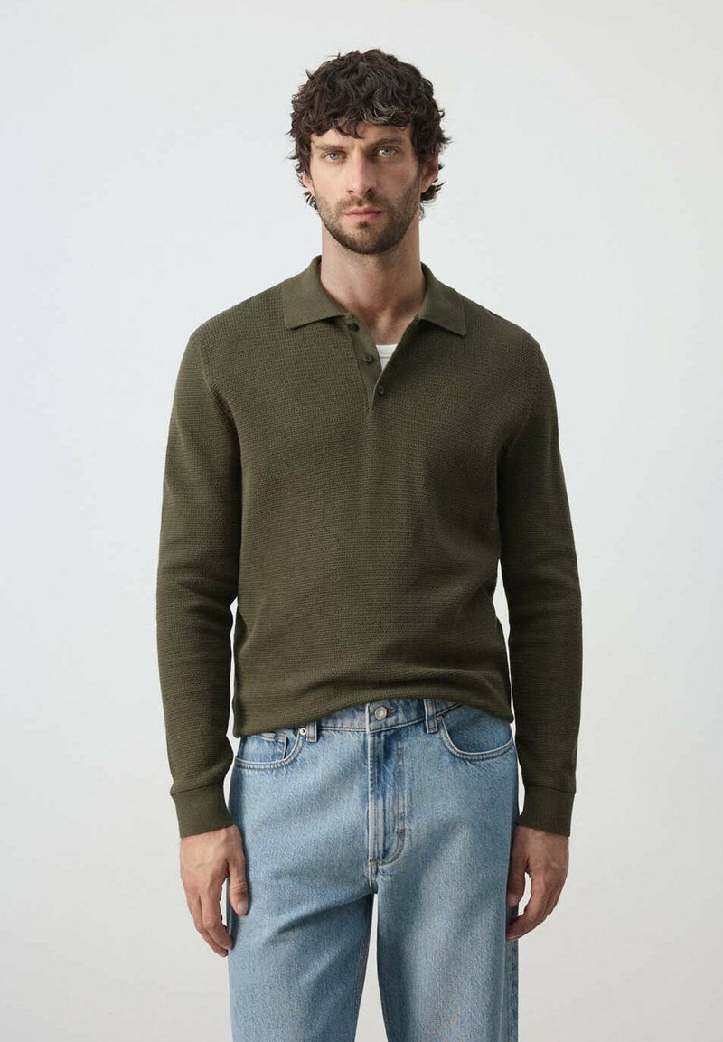 Homme aux cheveux bouclés et foncés portant un polo manches longues vert olive et un jean bleu clair, debout devant un fond uni.