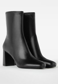 Schwarze Leder-Ankle-Boots mit sleekem Design, quadratischer Spitze und Blockabsatz, die sichtbare Nähte und eine glatte Textur aufweisen.
