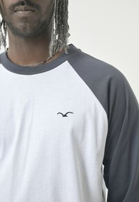 Graues und weißes Langarmshirt mit Raglan-Design, ausgestattet mit einem kleinen schwarzen bestickten Vogel-Logo auf der Brust. Weiche Stofftextur.
