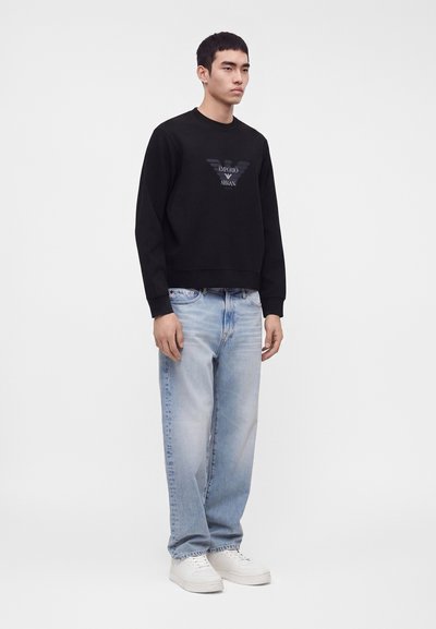 Άνδρας όρθιος με μαύρο φούτερ Emporio Armani, ανοιχτό μπλε τζιν και λευκά αθλητικά παπούτσια μπροστά σε λιτό ανοιχτό φόντο.