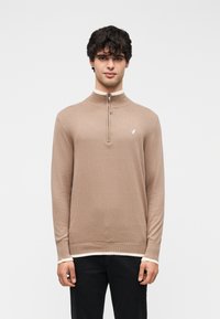 Pull en tricot marron à demi-fermeture éclair avec un col haut, un ourlet côtelé et des poignets crème contrastants. Petit logo sur la poitrine gauche.