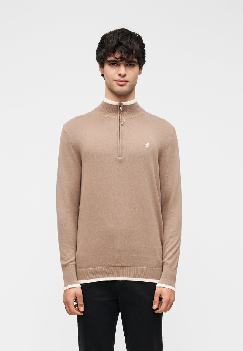 Pull en tricot marron à demi-fermeture éclair avec un col haut, un ourlet côtelé et des poignets crème contrastants. Petit logo sur la poitrine gauche.