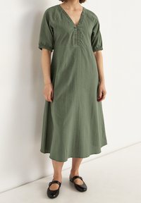 Oliwkowa sukienka midi z teksturowanej tkaniny, dekoltem w szpic i krótkimi bufkami, zestawiona z czarnymi butami Mary Jane.