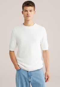 Jonge man met kort haar, gekleed in een wit fijngebreide korte mouw trui en lichtblauwe spijkerbroek, staand met één hand in zijn zak.