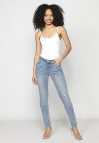 Jeans ajustados de color azul con detalles turquesa adornados en los bolsillos, lavado claro y ligeras desgastaciones en las rodillas, combinados con una camiseta sin mangas blanca.