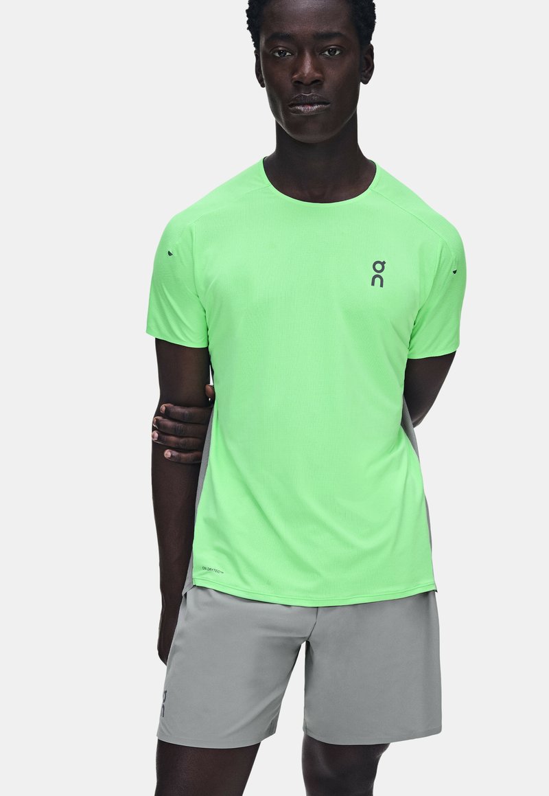 Homme portant un t-shirt de sport vert fluo à manches courtes et un short gris clair, debout avec un bras plié et la main reposant sur le bras opposé.
